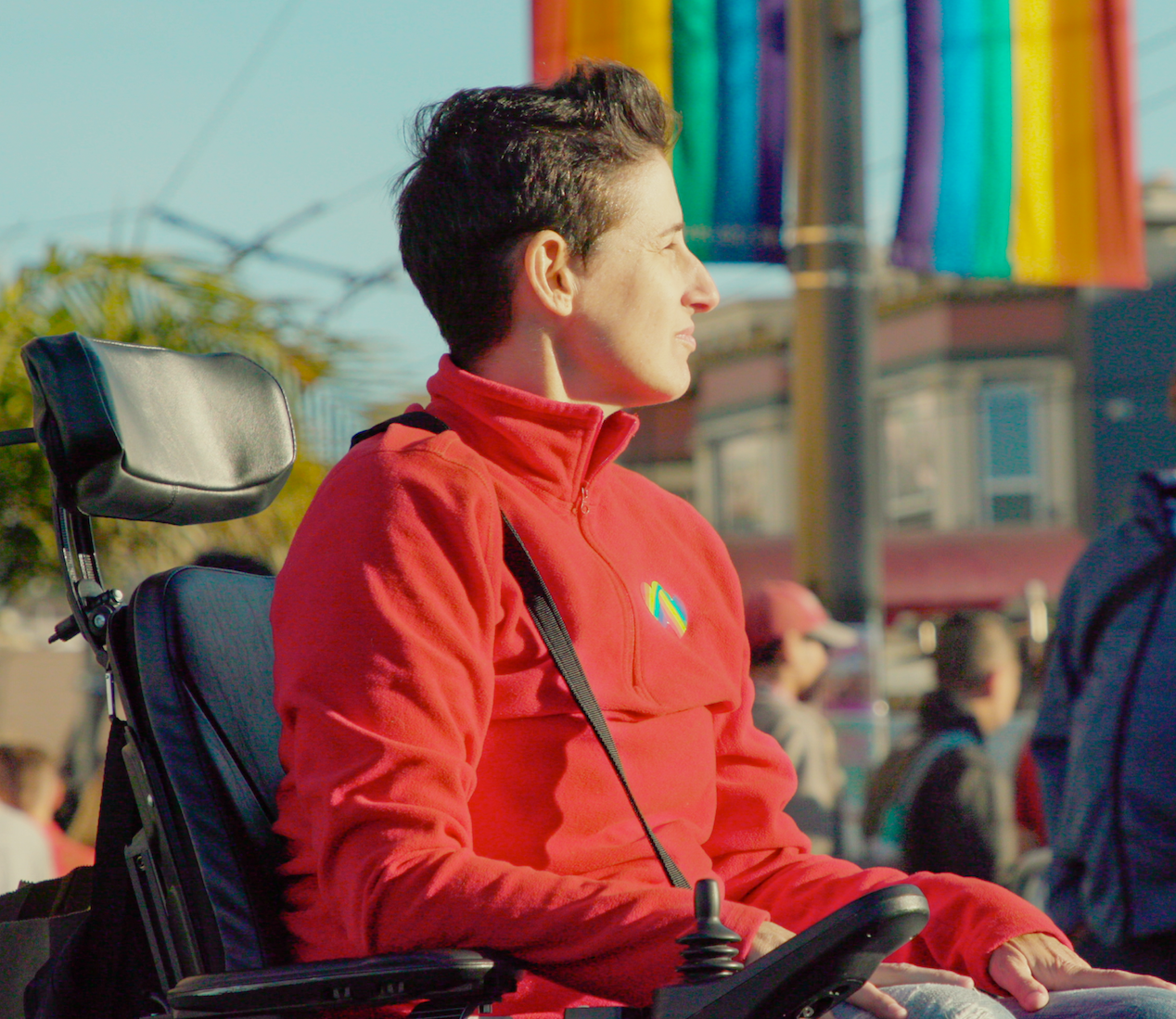 Franco Stevens sitting in their wheelchair outdoors, looking ahead pensively, with the Pride flag behind them.