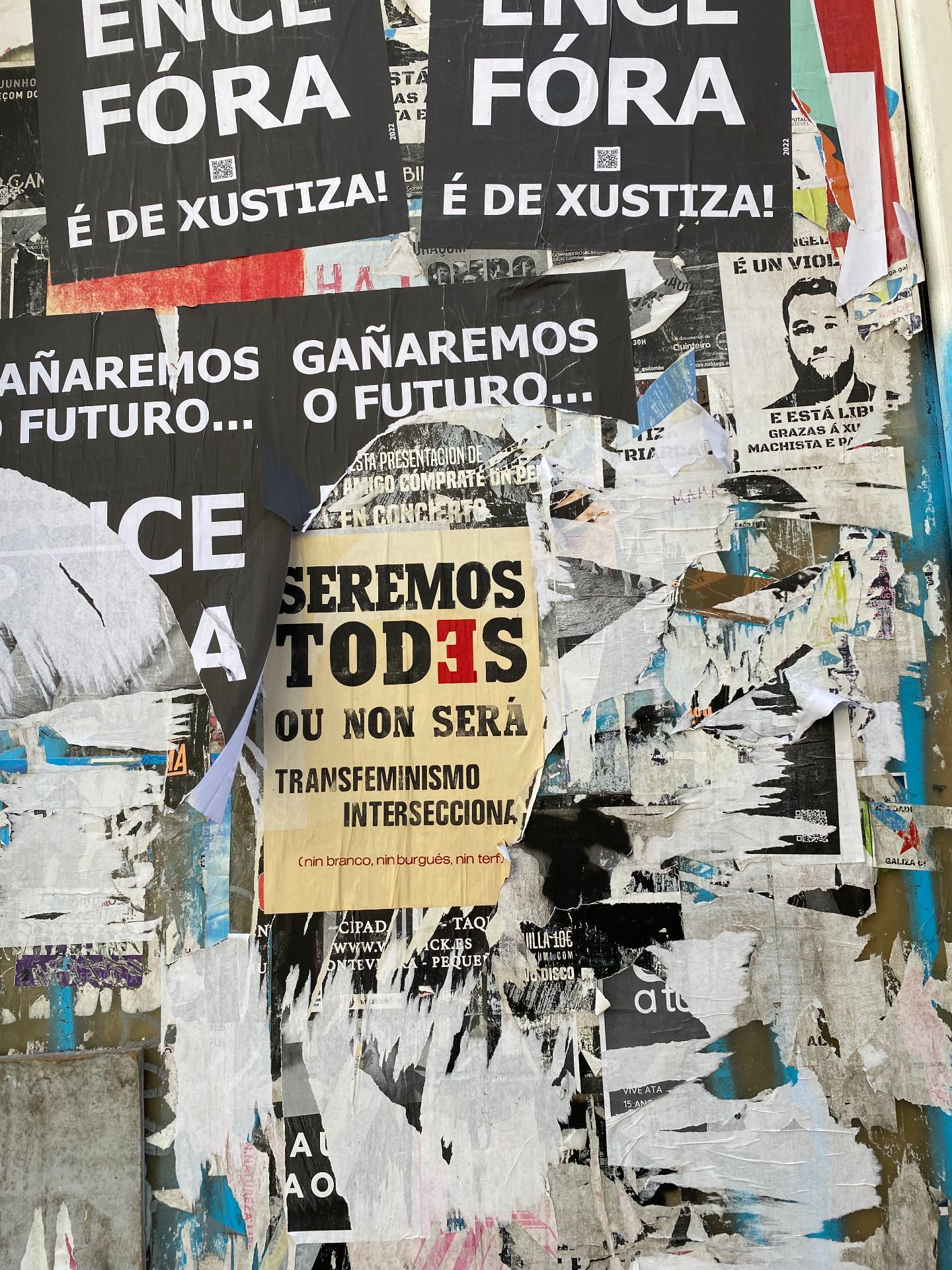 Foto de una pared con distintos carteles, algunos medio arrancados, y en el centro uno en el que se leer "Seremos todes ou non será. Transfeminismo intersaccional (nin binarrio, nin burgués, nin terf)".