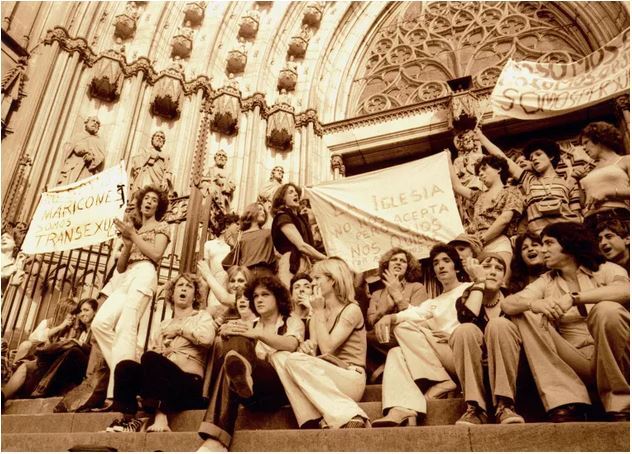 Fotografía en sepia de personas trans en la escalinata de una iglesia con pancartas en las que se lee "No somos maricones, somo transexuales", "La Iglesia no nos acepta, pero Dios nos quiere" y "No somos (algo), no somos objetos, somos personas".