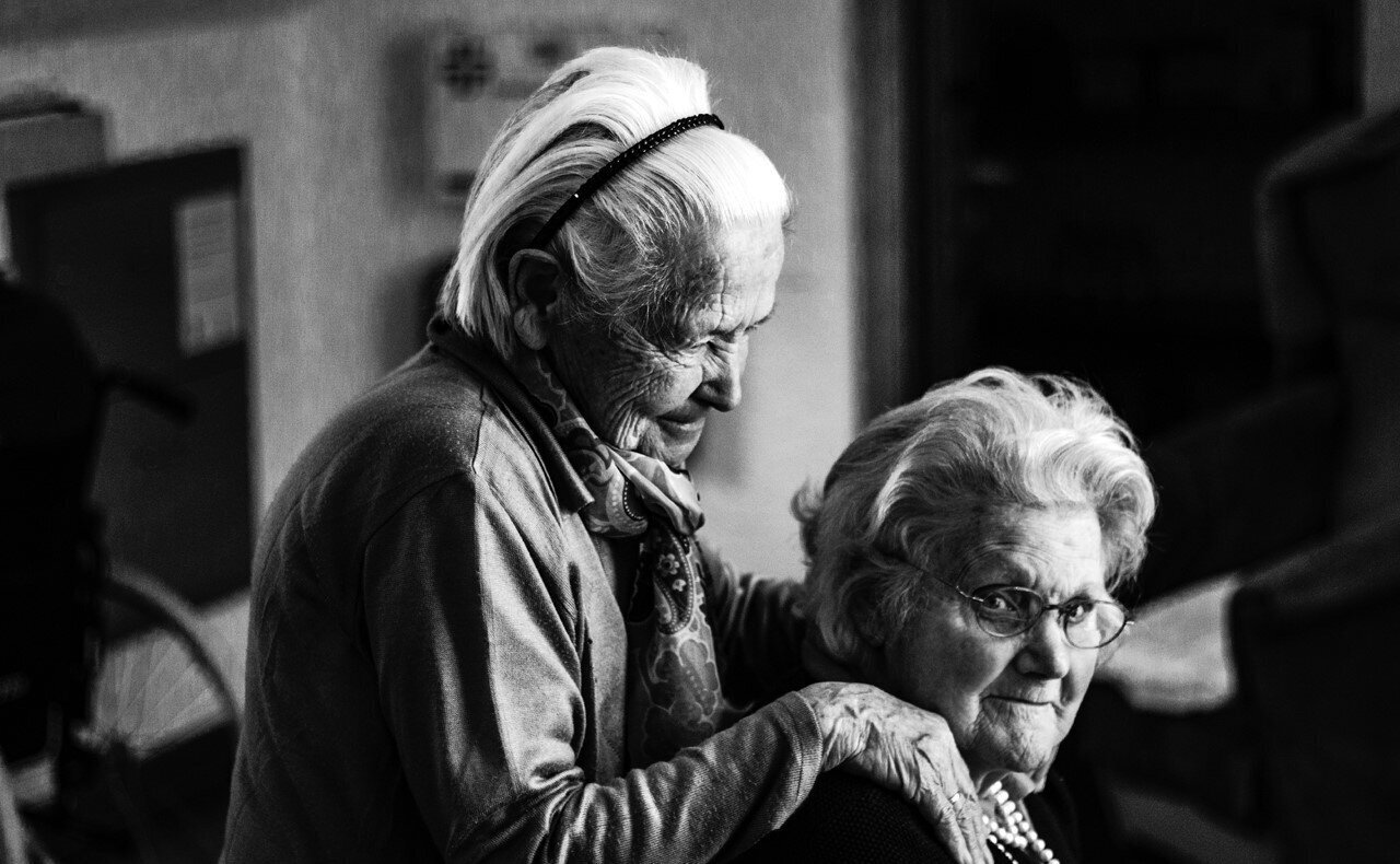 Foto ilustrativa de dos personas ancianas, una sentada, mirando al espectador, y otra de pie sujetando a la primera por los hombros