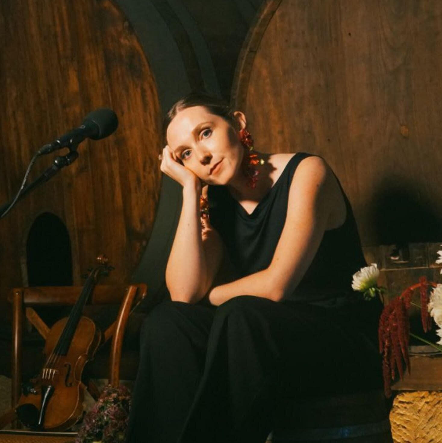 Fotograf&iacute;a de M&oacute;nica Morado, persona blanca que sale mirando a la c&aacute;mara en actitud pensativa y vestida de negro al uso de les int&eacute;rpretes de m&uacute;sica cl&aacute;sica. Delante de ella hay un micr&oacute;dono, a la izquierda se ve un viol&iacute;n y a la derecha, unas flores.