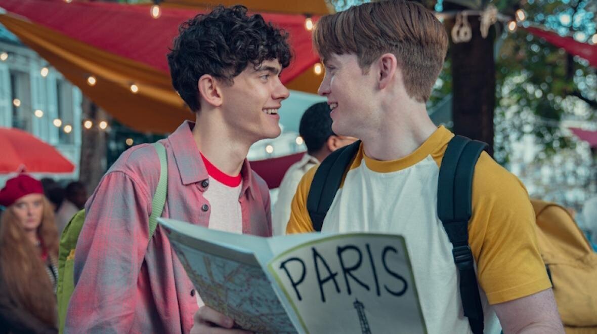 Fotograma de la segunda temporada de la serie Heartstopper en la que se ve a los protagonistas, Nick y Charlie, sonri&eacute;ndose el uno al otro mientras sujetan la gu&iacute;a de su viaje escolar a Par&iacute;s.