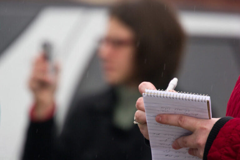 Fotografia con una persona al fondo, muy desenfocada, que est&aacute; grabando algo con su tel&eacute;fono m&oacute;vil. En primer plano, las manos de otra persona tomando notas en una libreta.