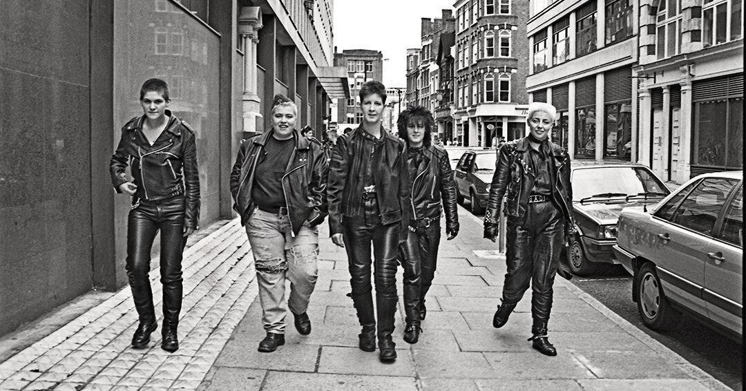 Fotografía de un paisaje urbano, en una acera junto a un edificio alto y coches aparcados al otro lado. En el centro, un grupo de personas queer de pelo corto, vestides con chaquetas y pantalones de cuero o vaqueros rotos.