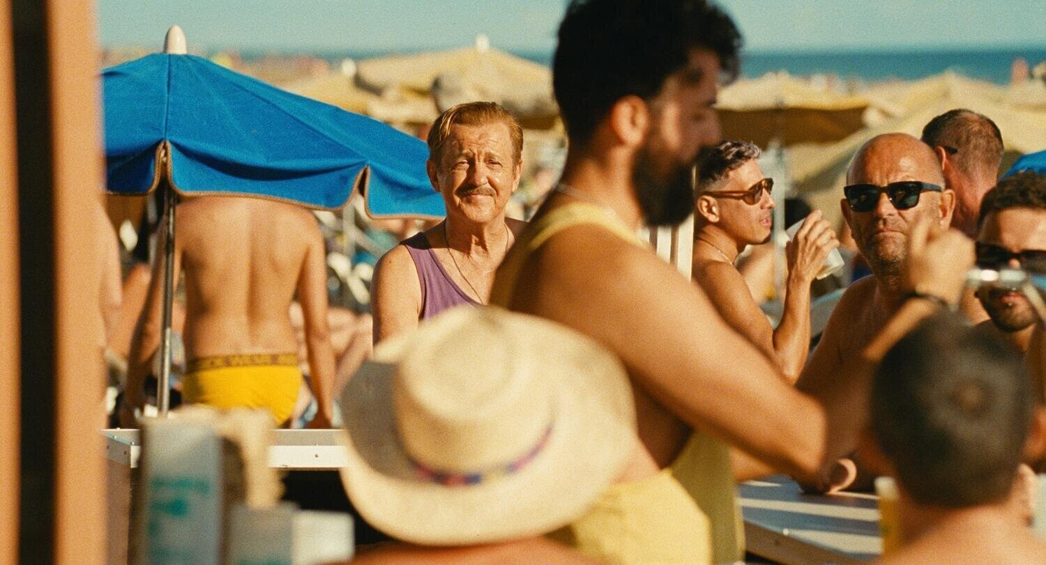 Fotograma de la película Maspalomas en la que se ve al protagonista en segundo plano en la playa, entre un montón de gente de distintas edades.