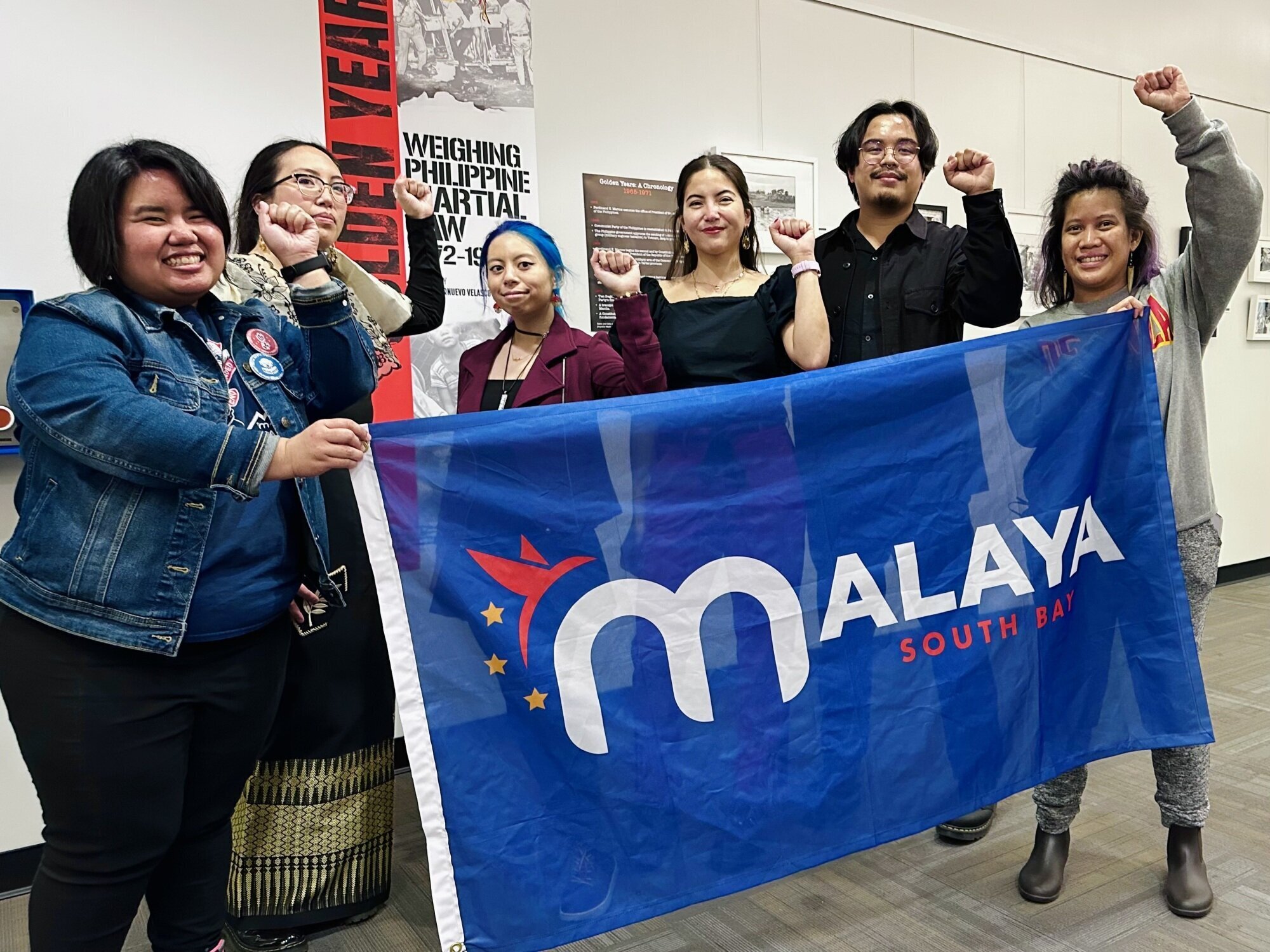 6 people with their left fist in the air, they are holding up a banner that says Malaya South Bay