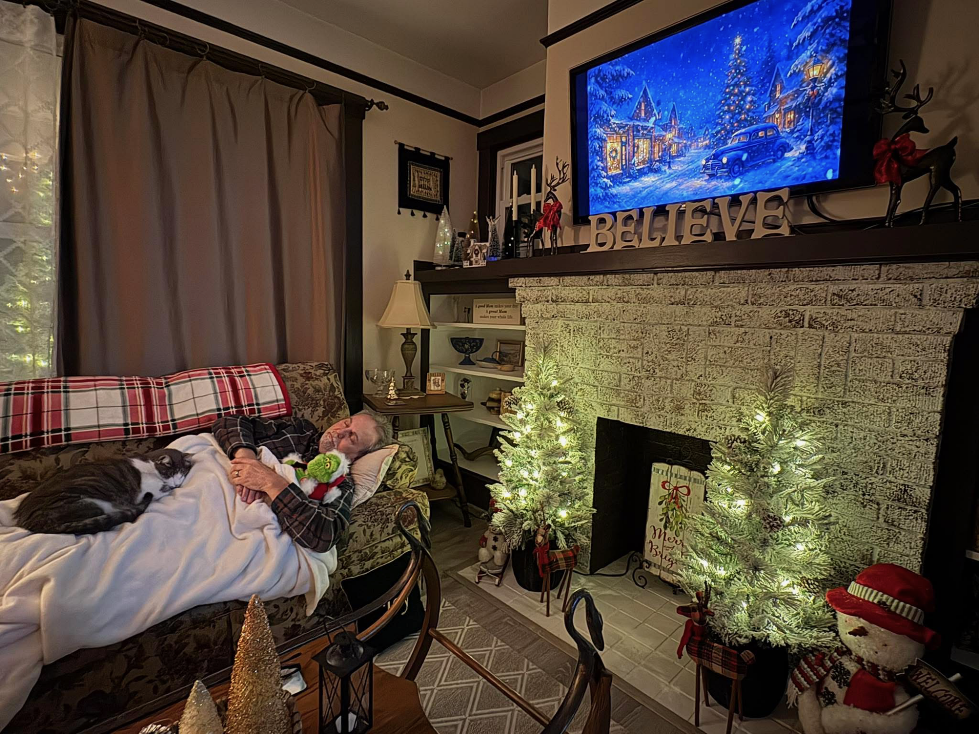 man sleeping on a couch with a plush Grinch and a cat sleeping on his lap, in a living room decorated for Christmas