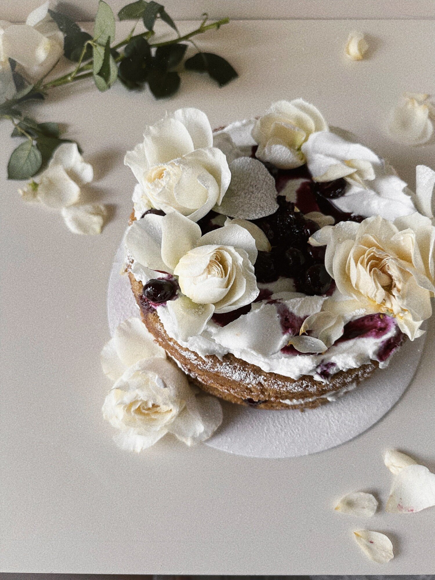 An overhead shot shows an earl grey olive oil cake with blueberry lavender compote and coconut whipped cream. The cake is adorned with white roses and petals.