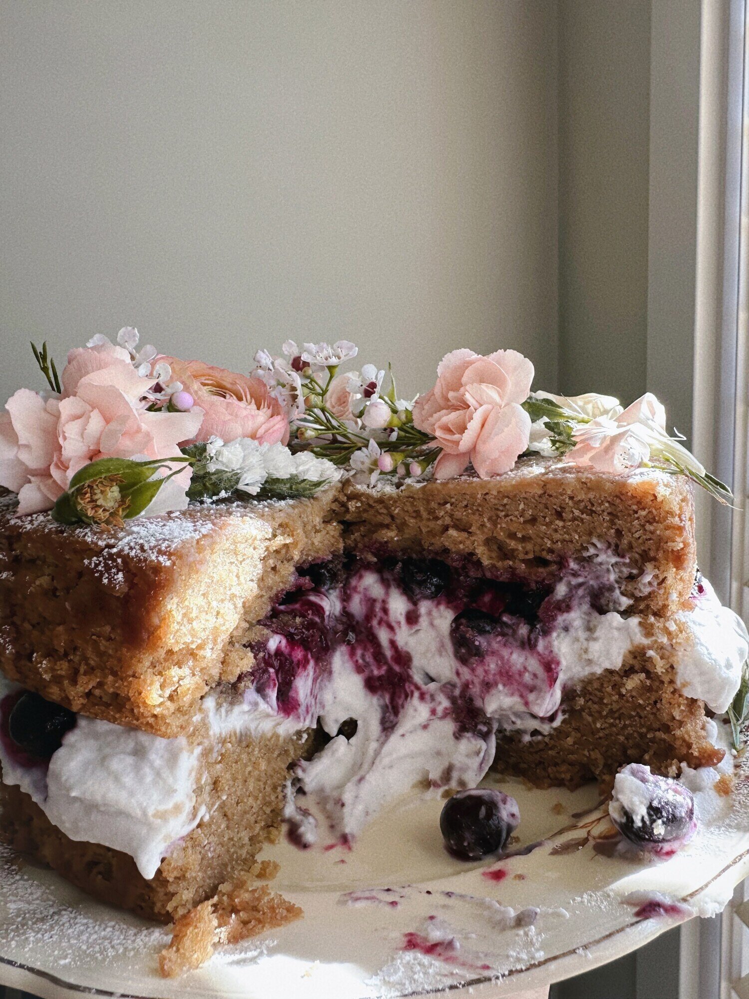 A head-on shot shows an earl grey olive oil cake with blueberry lavender compote and coconut whipped cream. A couple slices have been removed showing the lush interior of the cake and its layers. Flowers garnish the top.