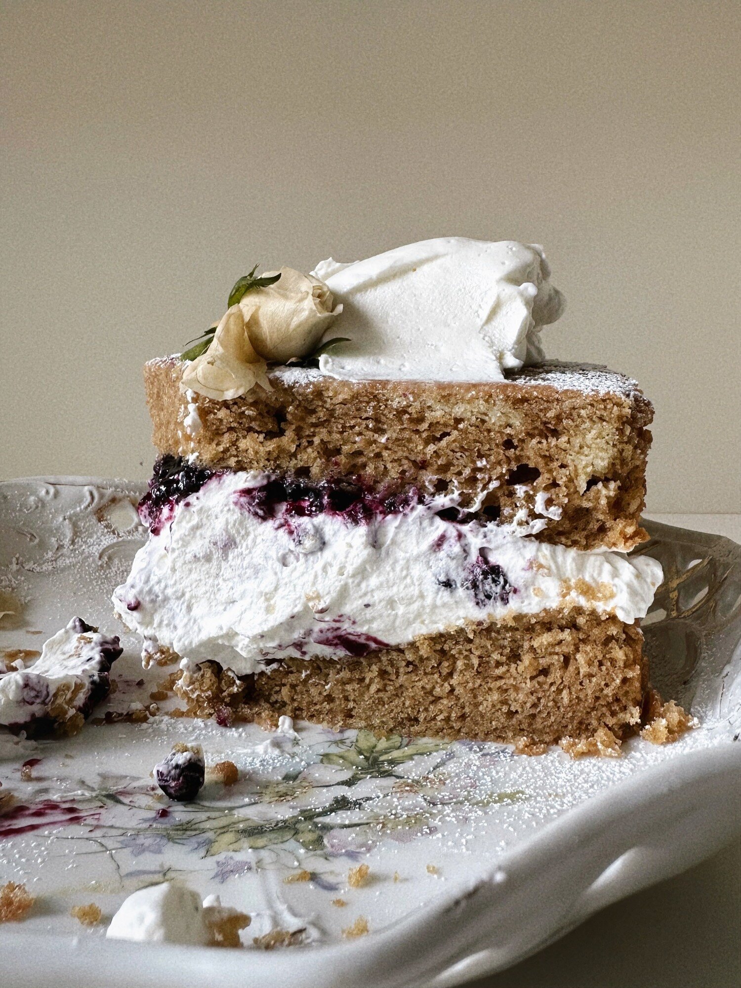 A head-on shot shows a layered slice of earl grey olive oil cake with blueberry lavender compote and coconut whipped cream. The cake is garnished with a white rose.