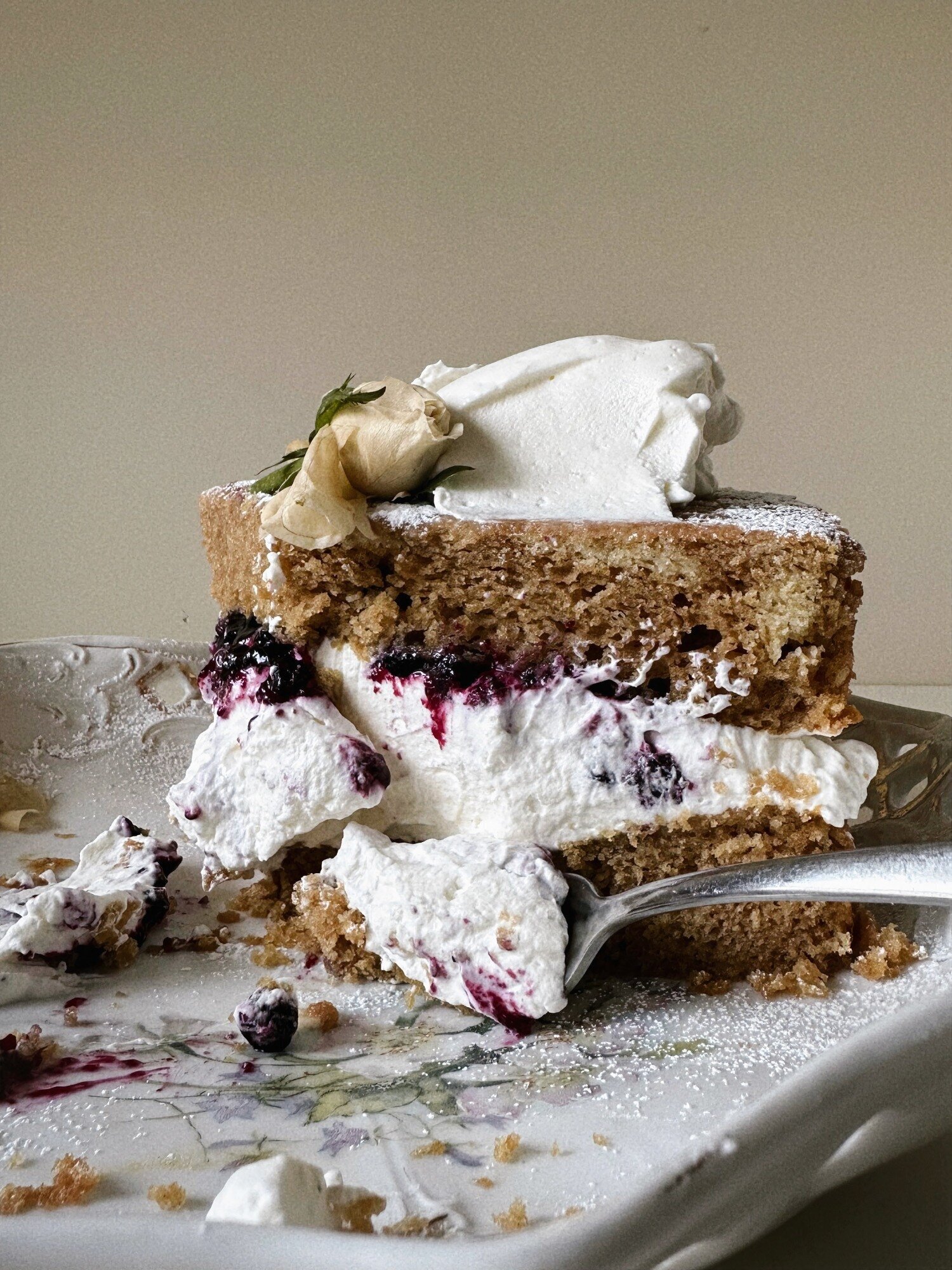 A head-on shot shows a layered slice of earl grey olive oil cake with blueberry lavender compote and coconut whipped cream. A fork has removed a bite and the cake is garnished with a white rose.